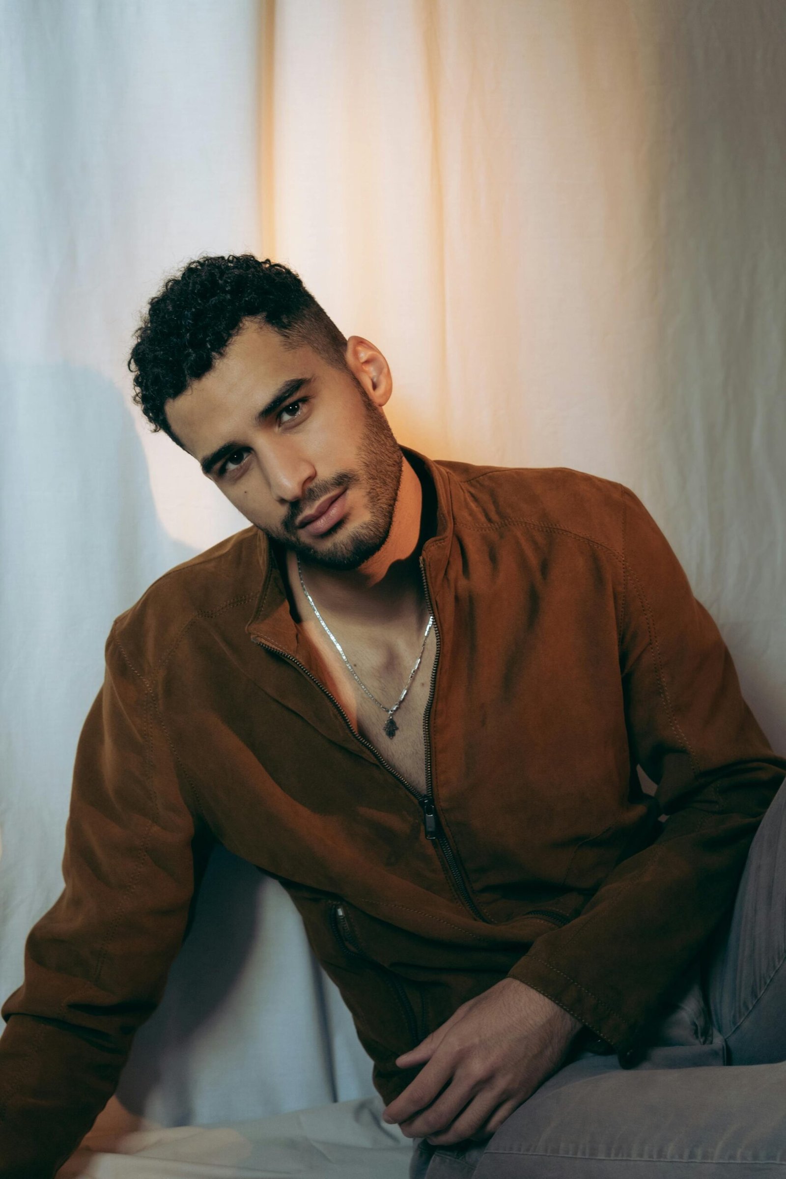 Elegant and stylish portrait of a man in a brown jacket sitting against a neutral backdrop.